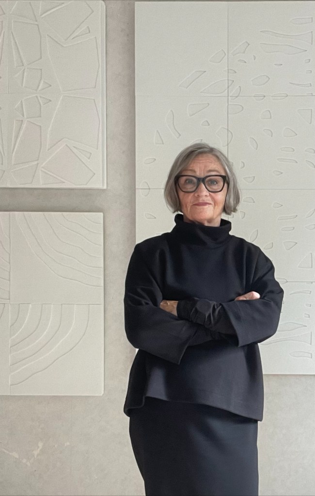 Carola Wiklund, konstnär och designer, är klädd i svart och står med armarna i kors framför akustikkonstverk i off-white.